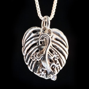 May include: A silver pendant necklace featuring a detailed gecko lizard climbing a stylized monstera leaf.