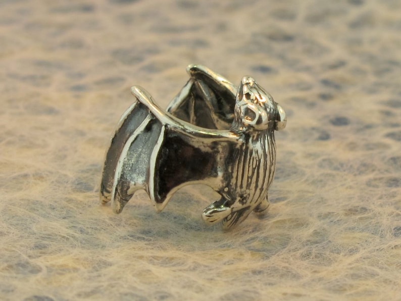 May include: A silver bat ring with detailed wings and a textured body. The ring is designed to be worn on the finger.