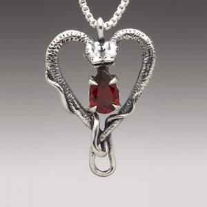 Op de afbeelding: Een zilveren ketting met een hanger met twee slangenhoofden die een hart vormen met een rode edelsteen in het midden.