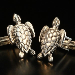 May include: A pair of silver cufflinks featuring a detailed turtle design. The turtles are facing forward with their heads raised and their legs outstretched. The cufflinks are attached to a silver bar.