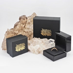 Op de afbeelding: Twee zwarte dozen met gouden letters die "Marty Magic" lezen en een kleinere zwarte doos met een gouden ring erin.