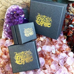 May include: Three black gift boxes with gold lettering that reads "Marry Magic" on each box. The boxes are sitting on a bed of purple and pink crystals.