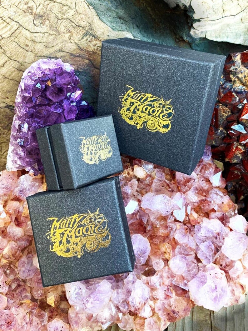 May include: Three black gift boxes with gold lettering that reads "Marry Magic" on each box. The boxes are sitting on a bed of purple and pink crystals.