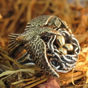 Könnte beinhalten: Ein silberner Ring, der einen Vogel darstellt, der ein Nest mit goldenen Eiern trägt. Der Ring ist mit strukturierten Federn und einem geflochtenen Nest verziert.