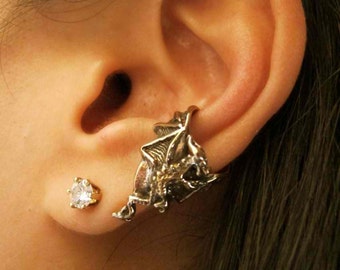 May include: A silver ear cuff in the shape of a bat with a small clear crystal stud earring in the lower lobe of the ear.