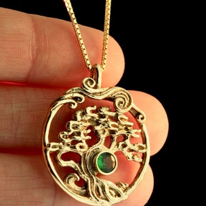 May include: A gold necklace with a pendant featuring a detailed tree of life design with a green gemstone at its center. The pendant is oval-shaped and has a delicate chain.