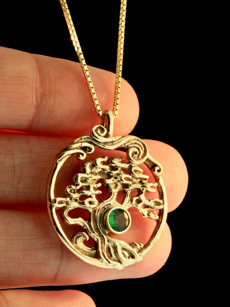 May include: A gold necklace with a pendant featuring a detailed tree of life design with a green gemstone at its center. The pendant is oval-shaped and has a delicate chain.