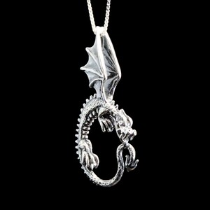 Op de afbeelding: Zilveren drakenhanger met uitgespreide vleugels, een gekrulde staart en gedetailleerde kenmerken. De hanger hangt aan een zilveren ketting.