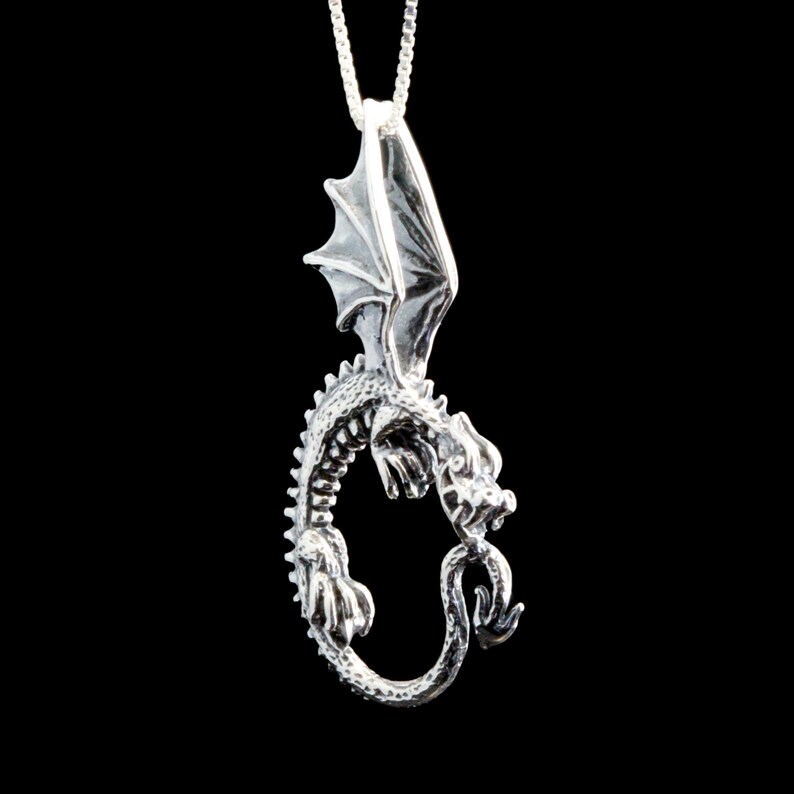 Op de afbeelding: Zilveren drakenhanger met uitgespreide vleugels, een gekrulde staart en gedetailleerde kenmerken. De hanger hangt aan een zilveren ketting.