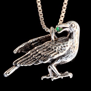 May include: A silver crow pendant with a green gemstone in its beak, hanging from a silver chain.