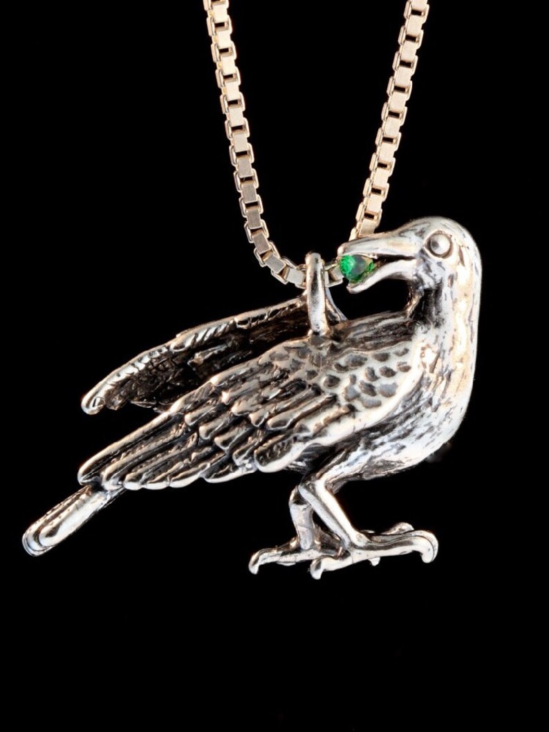 May include: A silver crow pendant with a green gemstone in its beak, hanging from a silver chain.