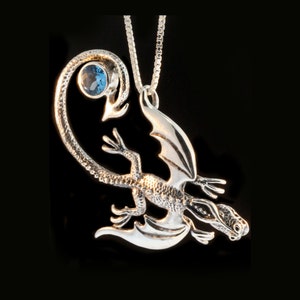 May include: Silver dragon pendant necklace with a blue gemstone. The dragon is curled up with its wings spread out. The necklace is on a silver chain.