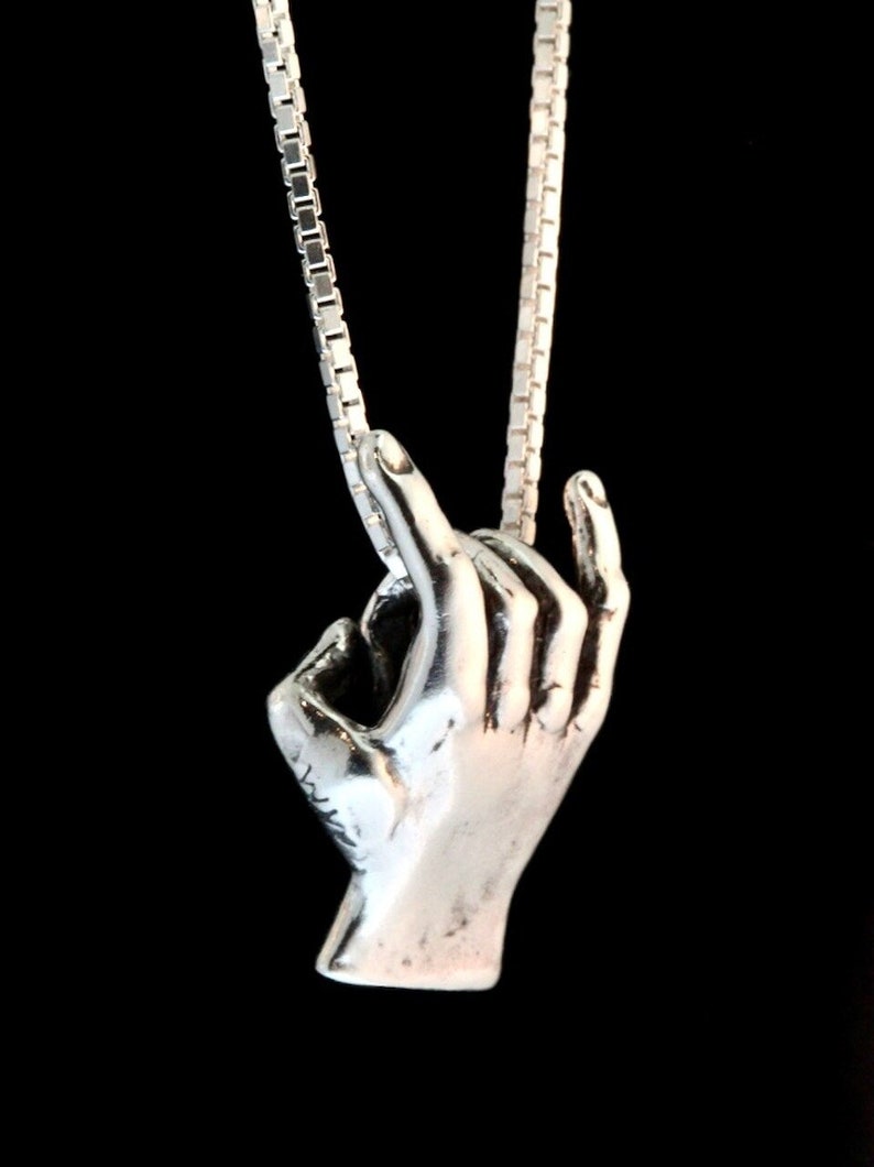 May include: A silver necklace with a hand pendant. The hand is making the 'okay' sign.