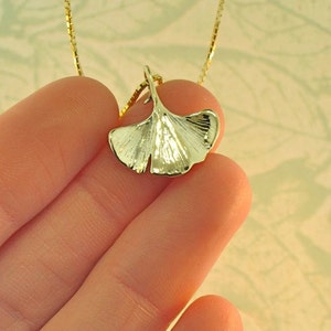 May include: A gold-toned necklace featuring a pendant shaped like a ginkgo leaf. The pendant has a smooth, reflective surface with subtle vein detailing and is suspended from a delicate, thin gold-toned box chain necklace.