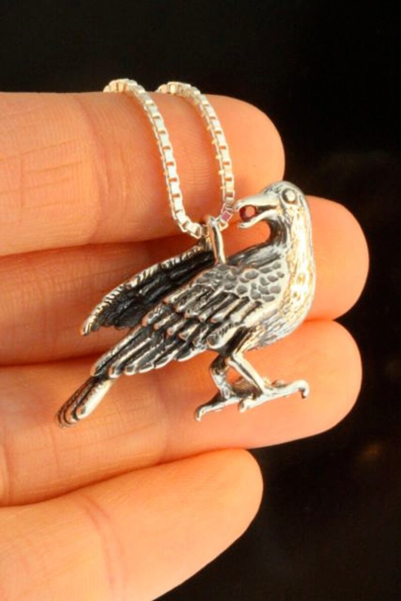 May include: A silver crow pendant with a red gemstone in its beak, hanging from a silver chain.