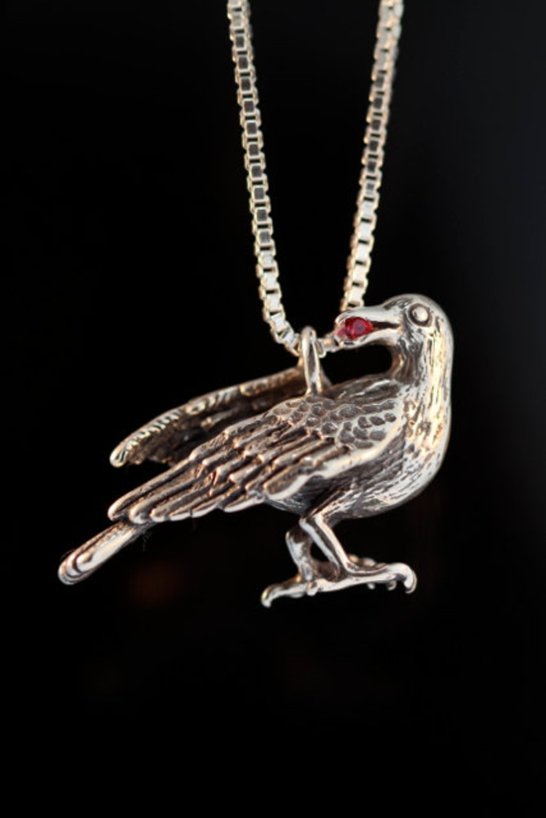 May include: A silver crow pendant with a red gemstone eye, hanging from a silver chain.