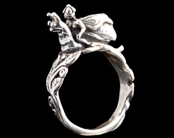 May include: A silver ring with a detailed design of a fairy riding a winged creature. The ring is made of metal and has a smooth finish.