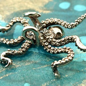 Puede incluir: Un amuleto de plata en forma de pulpo con tent&aacute;culos detallados. El amuleto est&aacute; hecho de metal y tiene una superficie texturizada.