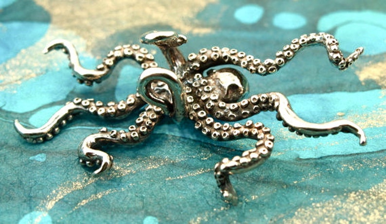 Puede incluir: Un amuleto de plata en forma de pulpo con tent&aacute;culos detallados. El amuleto est&aacute; hecho de metal y tiene una superficie texturizada.