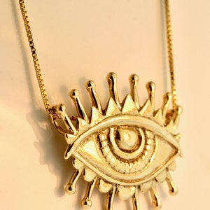 May include: A gold necklace with a large, detailed eye pendant. The eye has a sunburst design around it.