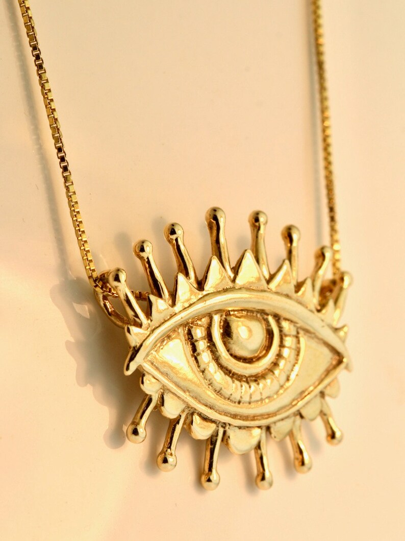 May include: A gold necklace with a large, detailed eye pendant. The eye has a sunburst design around it.