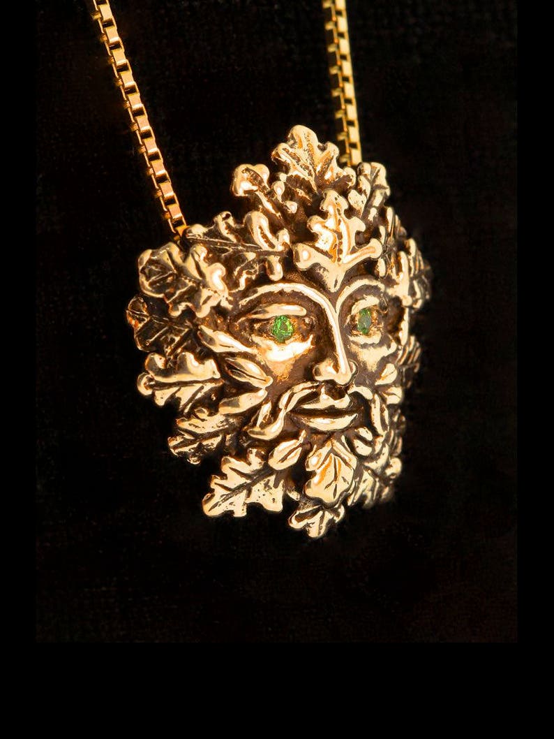Op de afbeelding: Een gouden ketting met een gedetailleerd Green Man-ontwerp met groene edelsteenogen. De hanger is omgeven door eikenbladeren.