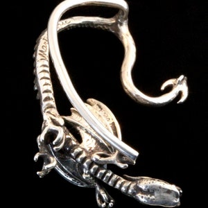 K&ouml;nnte beinhalten: Silberner Drachen-Ohrclip mit detailliertem Design. Der Clip ist offen und hat eine Drahtschlaufe zum Anbringen am Ohr.
