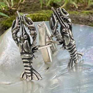 May include: A pair of silver-toned cufflinks, each shaped like a detailed lobster. The crustaceans are positioned on a pearlescent surface, with their claws raised. The cufflinks are connected by a rectangular bar.
