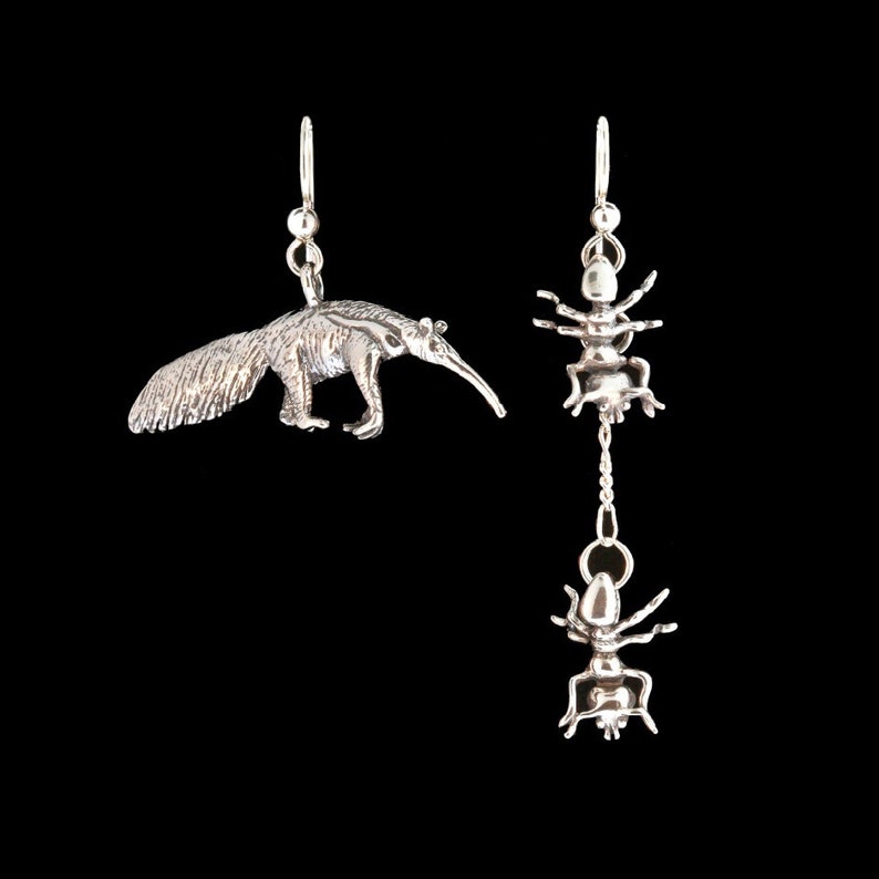 May include: A pair of silver earrings, one featuring an ant and the other an anteater. The ant earring has a dangling ant charm.