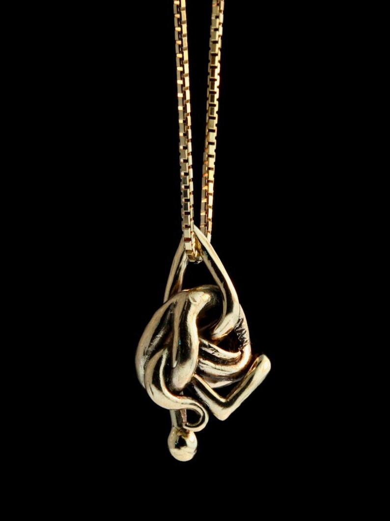 May include: A gold chain necklace with a pendant shaped like a stylized figure. The figure is abstract and appears to be in a contorted pose.