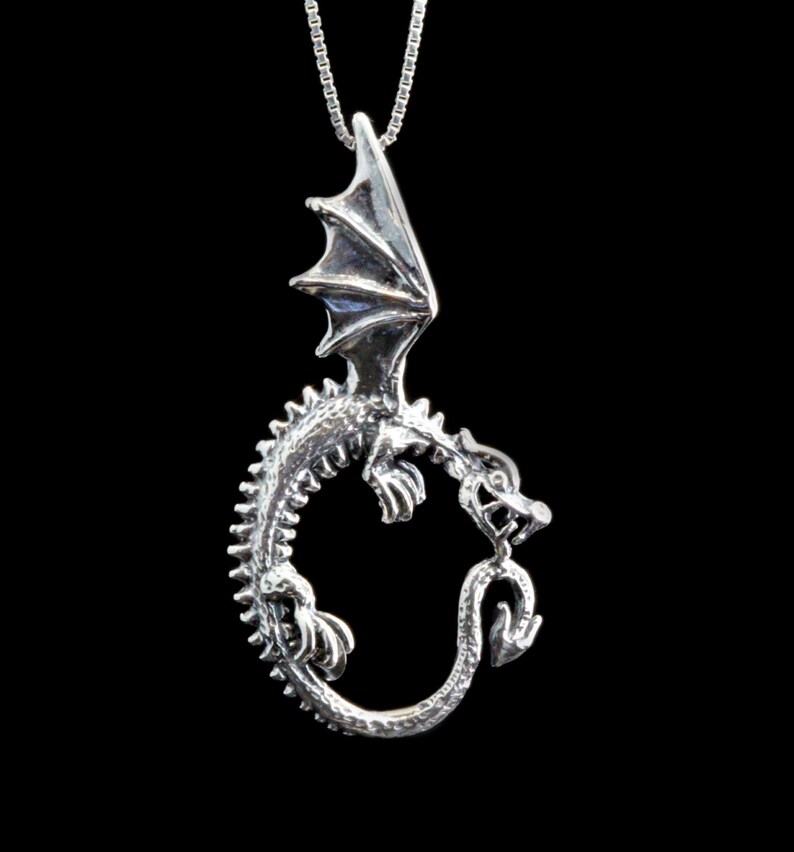 Op de afbeelding: Een zilveren drakenhangerketting met de draak opgerold in een cirkel en zijn vleugels uitgespreid. De draak is gedetailleerd met schubben en klauwen.