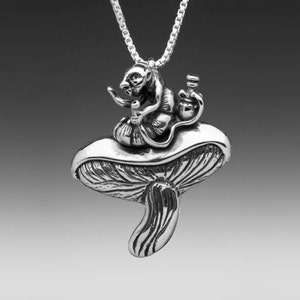 May include: A silver pendant necklace featuring a small monkey sitting on a mushroom. The monkey is holding a small bottle in one hand and a pipe in the other.