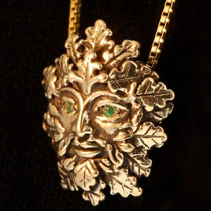 Op de afbeelding: Een gouden ketting met een gedetailleerd Green Man-ontwerp met smaragdgroene ogen. De Green Man is omringd door bladeren en takken, wat een natuur ge&iuml;nspireerd motief cre&euml;ert.
