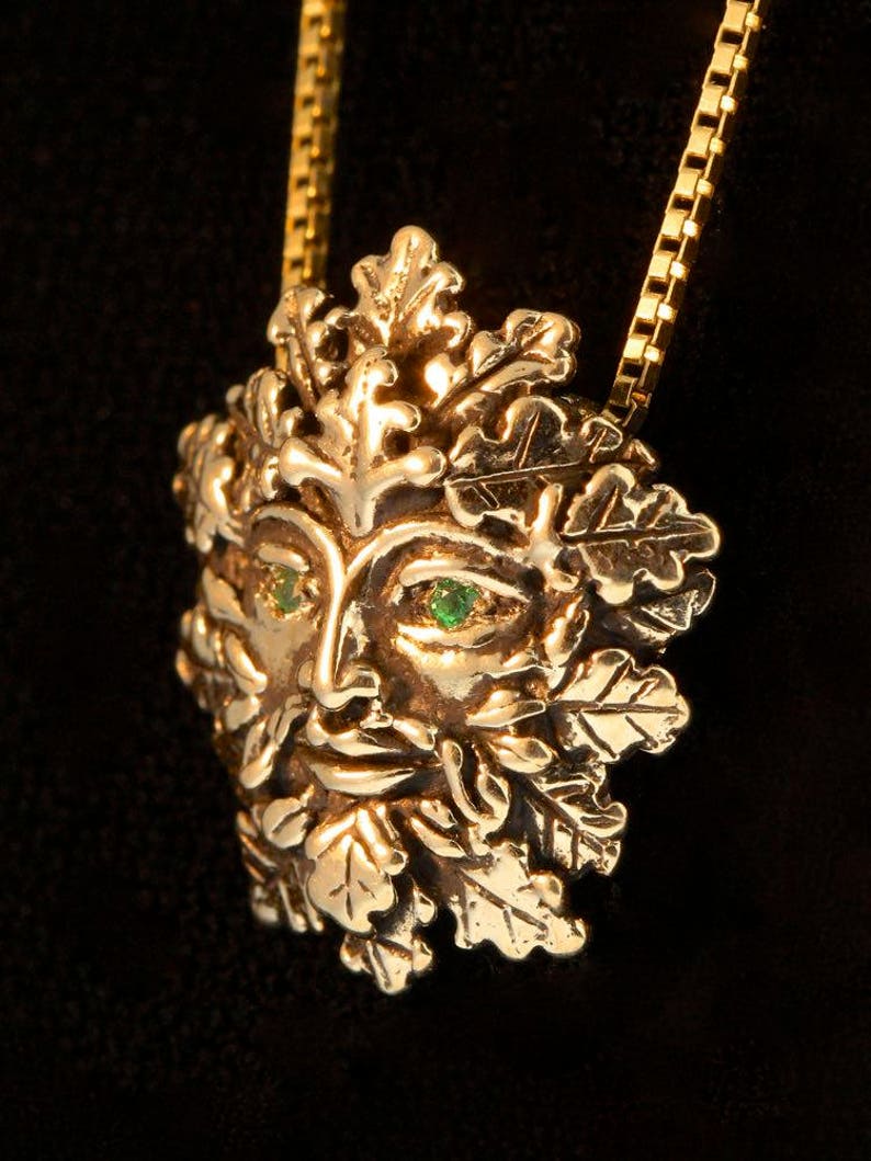 Op de afbeelding: Een gouden ketting met een gedetailleerd Green Man-ontwerp met smaragdgroene ogen. De Green Man is omringd door bladeren en takken, wat een natuur ge&iuml;nspireerd motief cre&euml;ert.