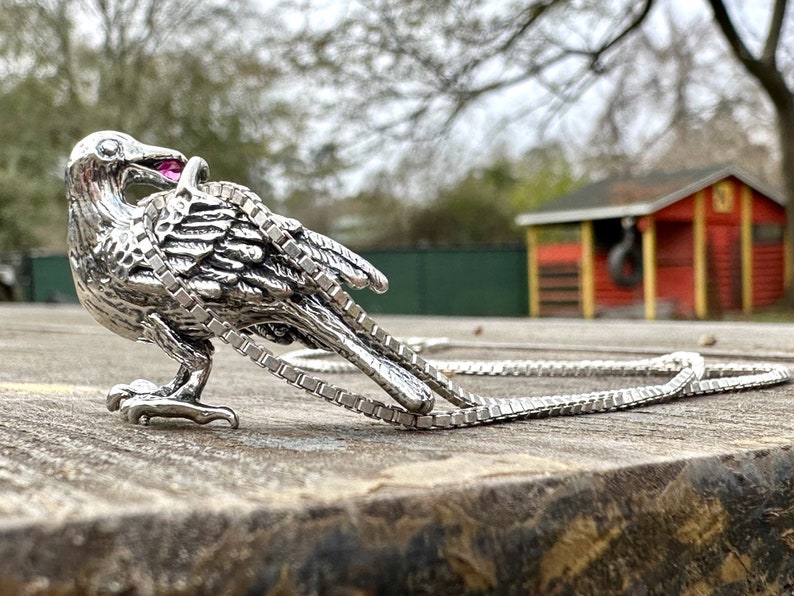 May include: A silver crow pendant with a pink gemstone in its beak, hanging on a silver chain.