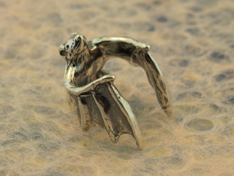 May include: A silver bat ring with wings spread out. The bat is facing forward with its mouth open.