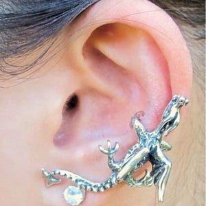 May include: A silver-toned ear cuff featuring a detailed lizard design. The lizard wraps around the ear, with a clear, round gemstone accent. The cuff is an alternative to traditional earrings.