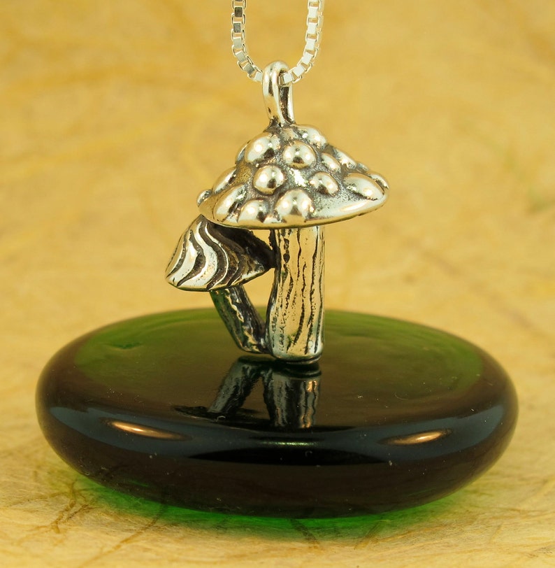 May include: A silver mushroom charm necklace with a detailed mushroom cap and stem. The charm is hanging from a silver chain and is displayed on a green glass surface.