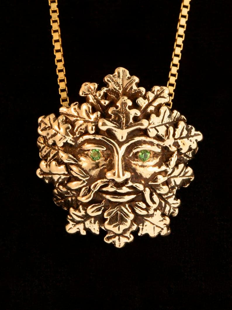 Op de afbeelding: Een gouden ketting met een gedetailleerd Green Man-ontwerp. Het gezicht van de Green Man is omringd door bladeren en heeft groene edelsteenogen.