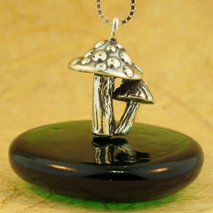 May include: A silver mushroom charm pendant with a detailed textured surface. The charm is hanging from a silver chain and is resting on a green glass surface.