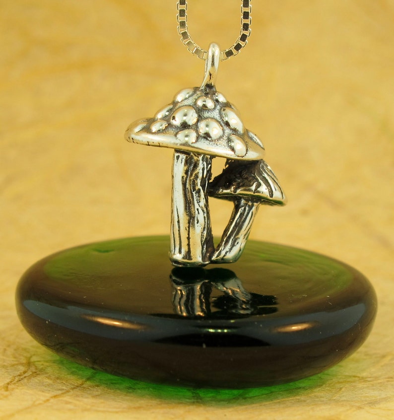 May include: A silver mushroom charm pendant with a detailed textured surface. The charm is hanging from a silver chain and is resting on a green glass surface.