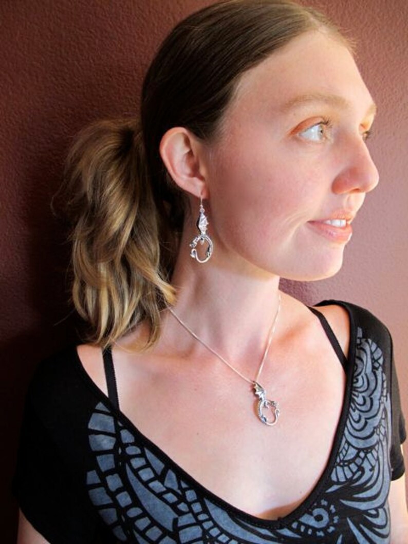 Op de afbeelding: Een vrouw draagt een zwart-grijs gem&ecirc;leerd shirt met een zilveren drakenhangerketting en bijpassende zilveren drakenoorbellen.