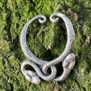 Pode incluir: Brinco de cobra de prata com duas cabeças de cobra que se olham. O brinco é texturizado e tem um acabamento martelado.