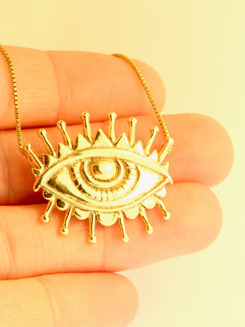 May include: A gold necklace with a large eye pendant. The eye is surrounded by a circle of rays.