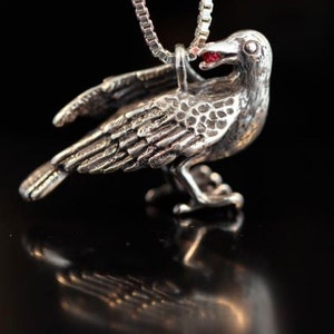 May include: Silver crow pendant necklace with a red gemstone eye. The crow is perched with its wings spread.