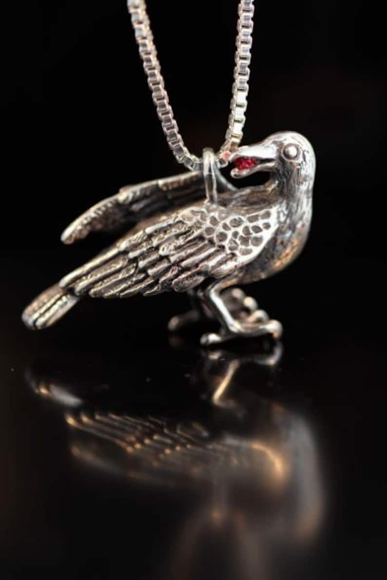 May include: Silver crow pendant necklace with a red gemstone eye. The crow is perched with its wings spread.