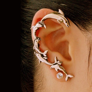May include: A gold-toned ear cuff featuring a design of multiple dolphins, with a single clear rhinestone stud earring below.
