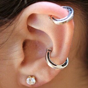 May include: A person's ear with two silver horseshoe-shaped earrings. The earrings are worn in the upper part of the ear, one above the other. A small, clear gemstone earring is worn in the lower lobe of the ear.