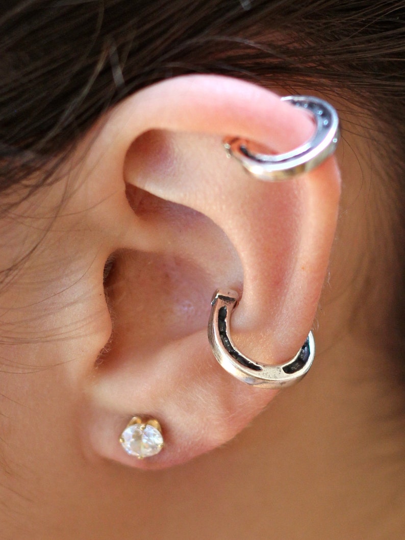 May include: A person's ear with two silver horseshoe-shaped earrings. The earrings are worn in the upper part of the ear, one above the other. A small, clear gemstone earring is worn in the lower lobe of the ear.