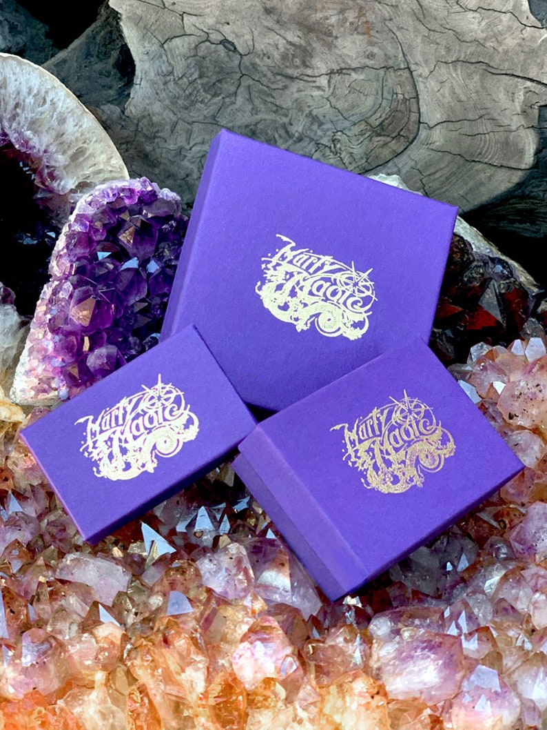 Puede incluir: Tres cajas moradas con letras doradas que dicen "Marry Magic" sobre un lecho de cristales de amatista.